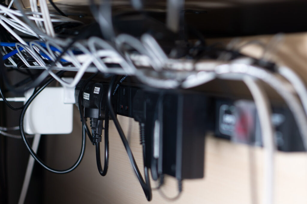 Under desk cable tray with lots of cables sits above a long server rack style multiway mains strip with many items connected.